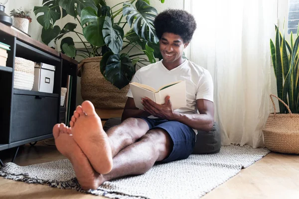 Glad, leende ung afrikansk amerikansk man läser en roman i mysiga vardagsrum. — Stockfoto