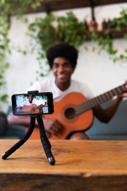 Smiling young african american man recording guitar online lesson with mobile phone. Selective focus on phone. Vertical.