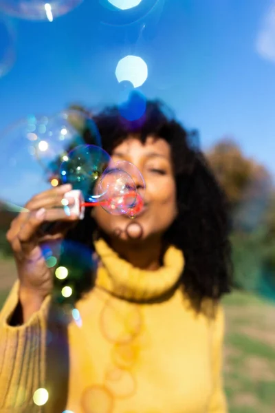 Vertikalt porträtt av ung afrikansk amerikansk kvinna som gör såpbubblor utomhus. Selektivt fokus på bubblor. — Stockfoto