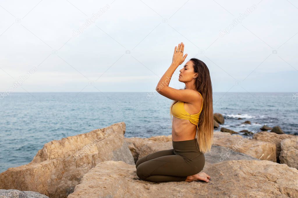 Young caucasian woman kneeling on a rock with hands in prayer by the ocean. Meditating in nature. Copy space.