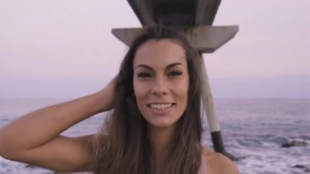 Mujer joven feliz mirando a la cámara tocándose el pelo en la playa al atardecer. — Vídeos de Stock