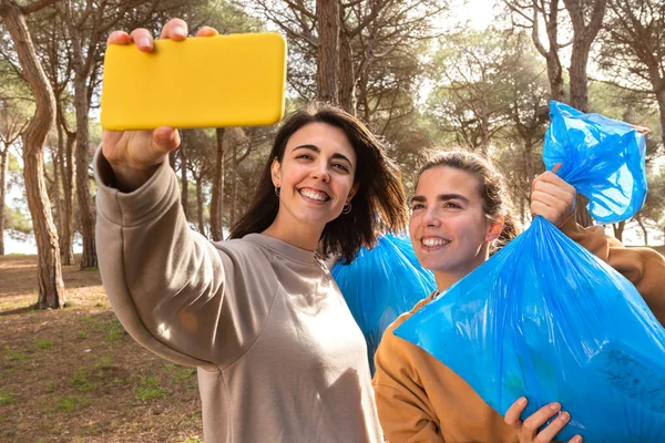 Unga kaukasiska kvinnor tar selfie med fulla soppåsar efter rengöring av skogen. — Stockfoto