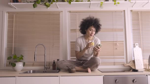 La donna di razza mista che fa colazione in cucina prende selfie e invia messaggi con il telefono mentre beve succo d'arancia. 4k — Video Stock