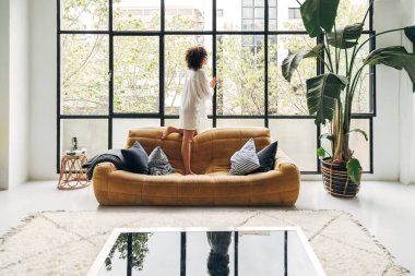 Genç Afro-Amerikalı kadın evdeki kanepede durmuş pencereden dışarı bakıyor..