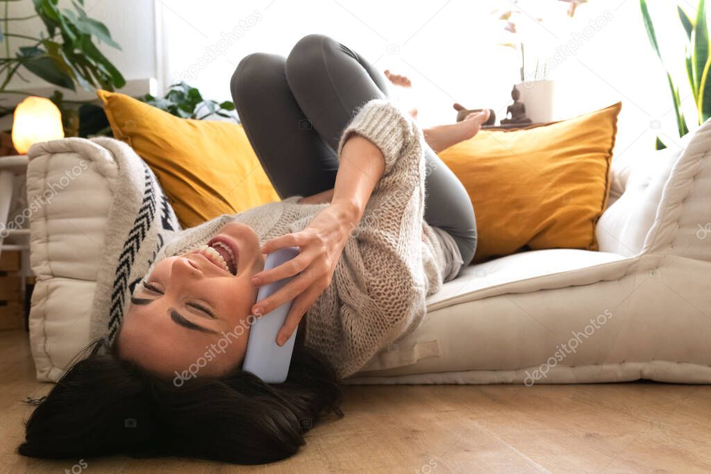Happy young caucasian woman lying upside down on the couch talking on cellphone laughing. Copy space.