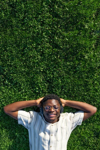 Draufsicht auf den entspannten schwarzen Mann. Glückliche schwarze Männchen, die im Gras liegen, hören Musik mit Kopfhörern. Kopierraum Vertikal — Stockfoto