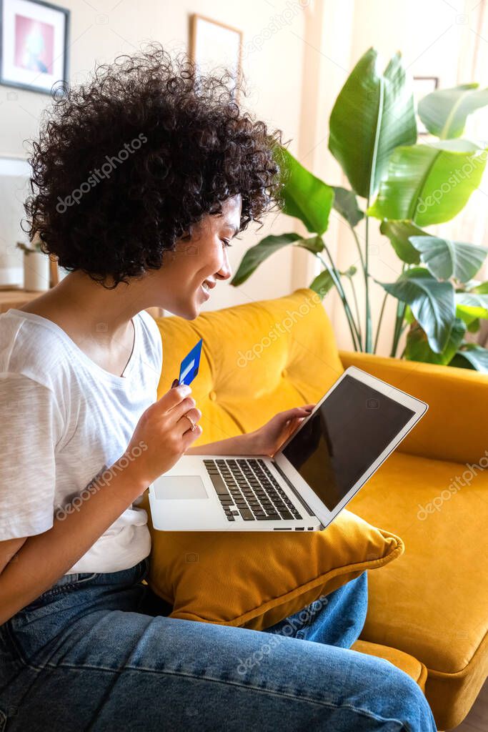 Side view of happy African american woman using credit card and laptop to shop online or order food at home sitting on sofa. Vertical image. Lifestyle and e-commerce concept.