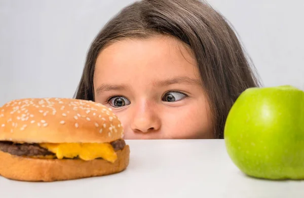 Çocuk Masanın Arkasından Dışarı Bakar Hamburgerle Elma Arasında Seçim Yapar — Stok fotoğraf