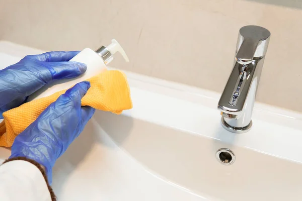 Kadın temizlikçinin banyo işleri yaparken, el sabunluğunu temizlerken. Mavi koruyucu eldiven giyiyor ve turuncu bir bez kullanıyor..