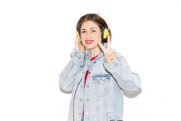 Beyaz arka planda yeşil kulaklıklarla müzik dinlerken gülümseyen neşeli ve güzel genç bir kadının portresi, zaferin olumlu bir jesti. Çok renkli günlük kıyafetler ve aksesuarlar..