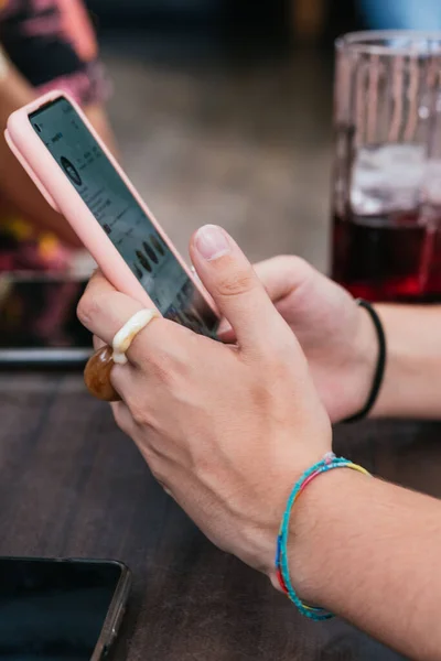 Hands of a fair-skinned young woman with finger rings and using an online application on a mobile phone, on the terrace of a bar with friends, holding a mobile phone.