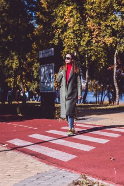 Kırmızı dizli, yeşil ceketli, kızıl saçlı bir kız güneşli bir günde sonbahar parkında yürüyor.