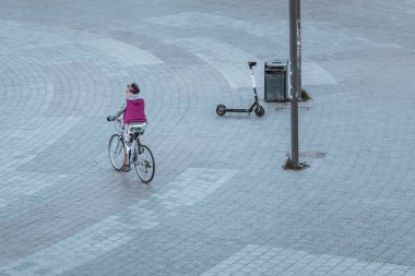 Kyiv, Ukrayna 26.10.2021: Mor yelekli kadın, sonbaharda boş gri bir meydanda elinde bir çöp tenekesi ve park etmiş bir scooterla bisiklet sürüyor.
