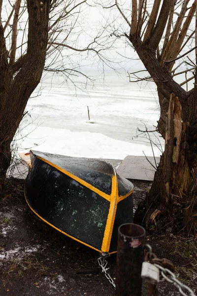 Altüst olmuş bir teknede donmuş bir gölün kıyısında, kasvetli ve bulutlu bir kış göğünün altında iki ağacın arasında uzanır.