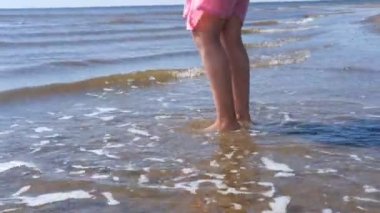 Bir kız kumda yalın ayak yürüyor. Deniz kıyısına su gelir, bir kız dalgalara atlar..