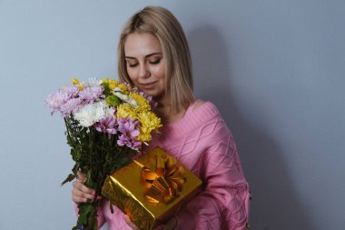 Altın bir kutuda bir hediye ve bir buket çiçek tutan bir kız. Hafif arka plan, seçici odaklanma. Kara Cuma, tatil, doğum günü, kadınlar günü kavramı.