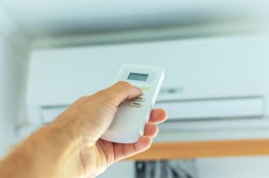 El uzaktan kumandayı kullanarak klimayı açar. Arka plandaki duvarda bir klima var.