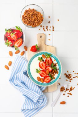 Kahvaltı gevreği ve beyaz masada taze çilek yapmak için malzemelerle dolu beyaz kasede Yunan yoğurdu. Üst görünüm