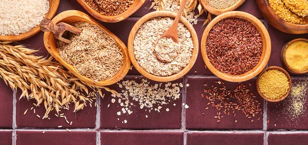 White, brown and red rice, buckwheat, millet, corn groats, quinoa and bulgur in wooden bowls on the light gray kitchen table. Gluten-free cereals. Top view with copyspace. Banner.