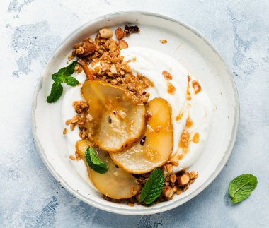 Gri seramik tabakta sağlıklı bir kahvaltı için karamelize armut, granola, fındık ve eritilmiş şeker içeren Yunan yoğurdu. Kırsal tarzda. Üst görünüm