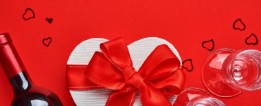 Kırmızı arka planda kırmızı kurdeleli, kalp şeklinde şarap, bardak ve hediye kutusu. Sevgililer Günü kartpostalı. Üst görünüm.
