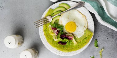 Kızarmış yumurta, avokado ve gri beton arka planda seramik tabakta salata yapraklı ıspanak yeşili krep. Sağlıklı kahvaltı kavramı. Seçici odaklanma. Üst Manzara. Boşluğu kopyala.