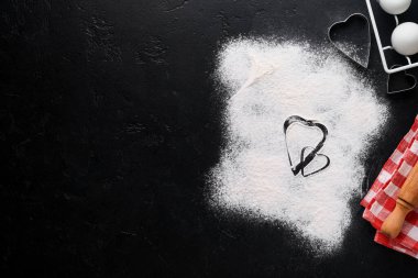 Sevgililer Günü 'nde arka planda kalp şekli, un, aletler ve siyah masada ev yapımı kurabiye pişirmek için malzemeler. Yemek arkaplanına bayılırım. Pankart. Üst görünüm.