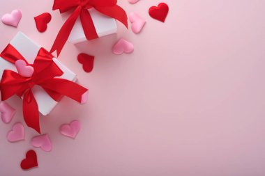 Kırmızı kurdeleli beyaz hediye kutuları ve pembe arka planda küçük kırmızı dekoratif kalpler. Kopyalama alanı olan üst görünüm. Sevgililer Günü ya da Düğün Romantik Konsepti. Kutlama kompozisyonu. Model.
