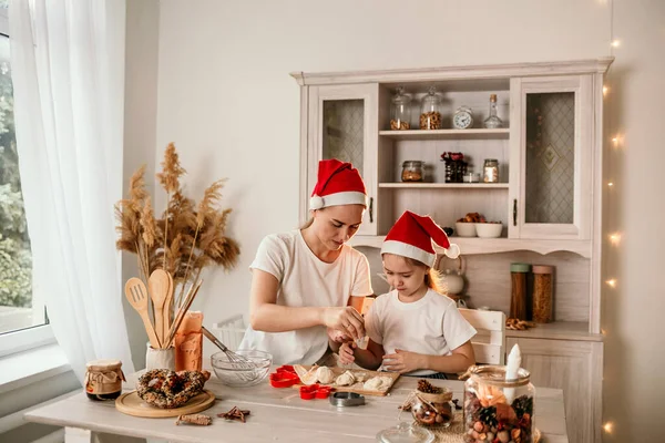クリスマスの帽子のお母さんと娘がキッチンに座って生地からクッキーを作っています — ストック写真