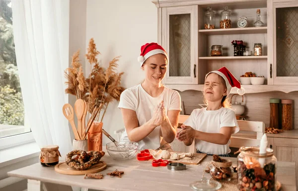 クリスマスの帽子の女の子とお母さんがテーブルに座って小麦粉で遊んでいます — ストック写真