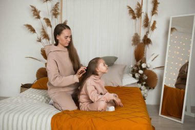 Anne yatağa oturur ve kızının saçını tarar.