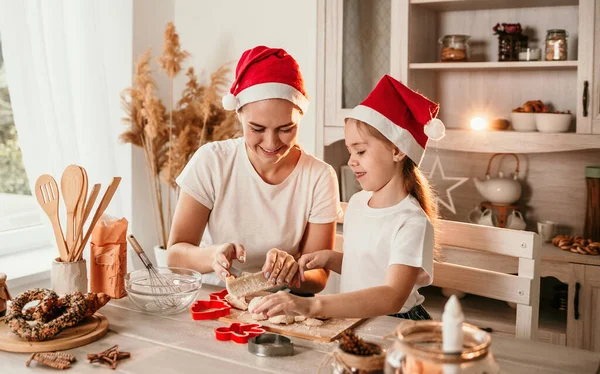 クリスマスの帽子の美しいお母さんと娘は台所のテーブルに座って生地をこねる — ストック写真