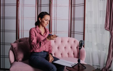 Pembe gömlekli bir kadın kanepede oturuyor ve telefonunu kullanarak online danışmanlık yapıyor.
