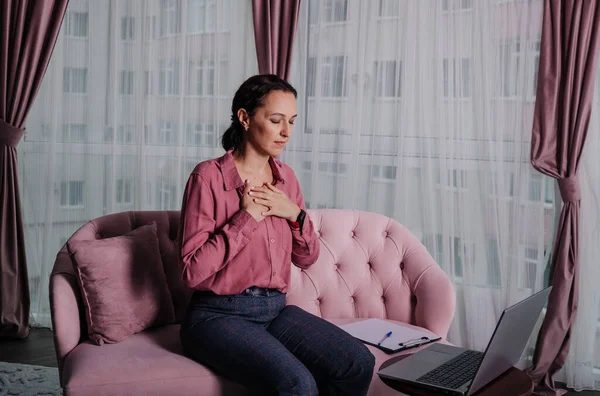 a woman in a pink shirt sits on the sofa and conducts a remote consultation using a laptop