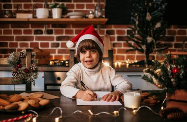 Beyaz örgü süveter ve kırmızı şapkalı küçük mutlu bir çocuk yeni yıl mektubu yazıyor.