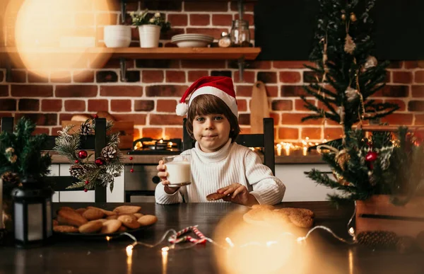 白いセーターを着た白人の男の子と新年の帽子はミルクとオートミールのクッキーでキッチンに座っています — ストック写真