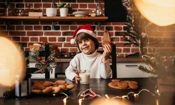 白いセーターを着た幸せな男の子と新年の帽子はミルクとオートミールのクッキーでキッチンに座っています — ストック写真