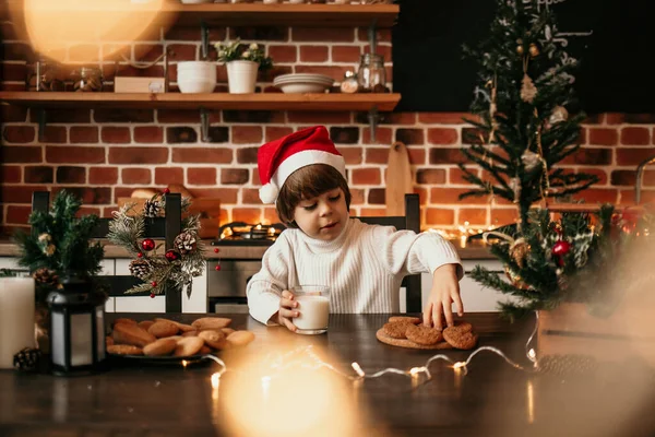 白いセーターを着た男の子と新年の帽子はミルクとオートミールのクッキーでキッチンテーブルに座っています — ストック写真
