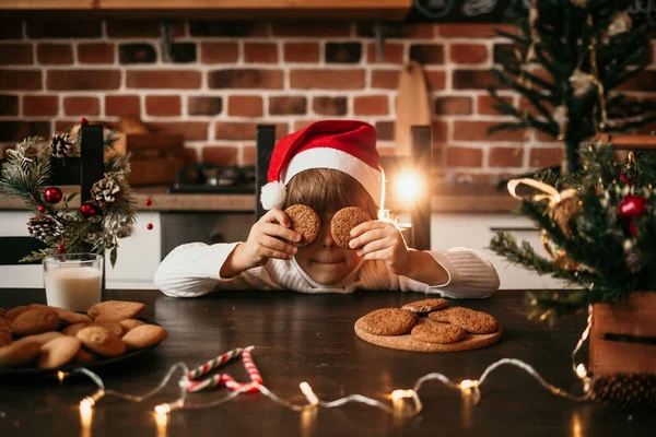白いセーターの男の子と新年の帽子がキッチンに座ってオートミールクッキーで目を閉じます — ストック写真