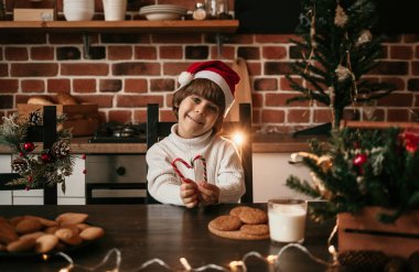 Beyaz örgü süveter ve yeni yıl şapkası giymiş küçük mutlu bir çocuk masada oturuyor ve çubukta lolipoplar tutuyor.