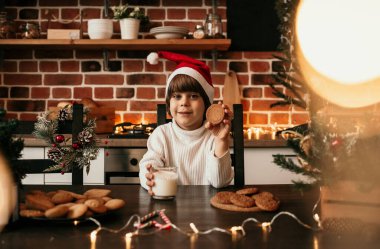 Beyaz örgü süveter ve yeni yıl şapkası takmış beyaz bir çocuk süt ve yulaf ezmeli kurabiyelerle dolu bir masada oturuyor.