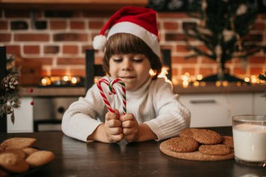 Yılbaşı şapkalı, masada oturan ve çubukta lolipop tutan bir çocuğun yakın çekimi. Lolipoplara odaklan.