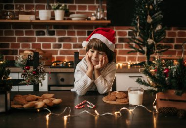 Beyaz örgü süveter ve yeni yıl şapkası giymiş rüya gören küçük bir çocuk mutfak masasında oturuyor.