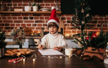 Beyaz örgü süveter ve kırmızı şapkalı küçük bir çocuk masada oturuyor ve yeni yıl mektubu yazıyor.
