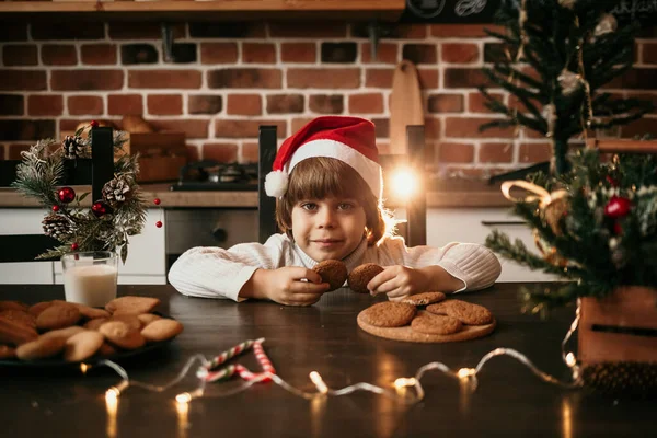 白いニットのセーターと新年の帽子の小さな男の子がテーブルに座ってオートミールクッキーを持っています — ストック写真