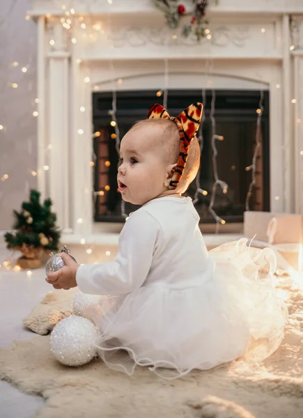 ふわふわのドレスを着た白人の赤ん坊と虎の耳を持つヘッドバンドは 新年の装飾が施された部屋の毛皮の敷物に座っています — ストック写真