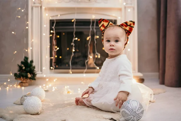ふわふわのドレスを着た赤ん坊の女の子と虎の耳を持つヘッドバンドは 新年の装飾が施された部屋の毛皮の敷物に座っています — ストック写真
