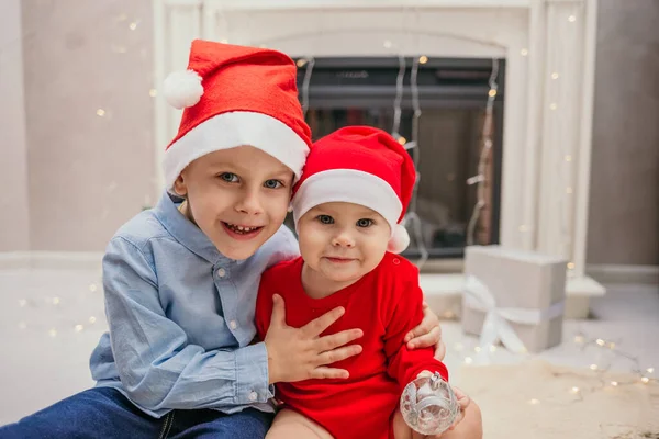 新年の赤い帽子の弟と妹が暖炉の近くの部屋に座っています — ストック写真