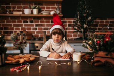 Beyaz örgü süveter ve kırmızı yeni yıl şapkası giyen beyaz bir çocuk mutfak masasında oturuyor ve yeni yıl mesajı yazıyor.