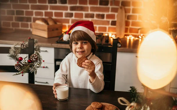 白いニットのセーターと赤いクリスマスの帽子のかわいい男の子がキッチンテーブルに座ってオートミールクッキーとミルクを食べています — ストック写真